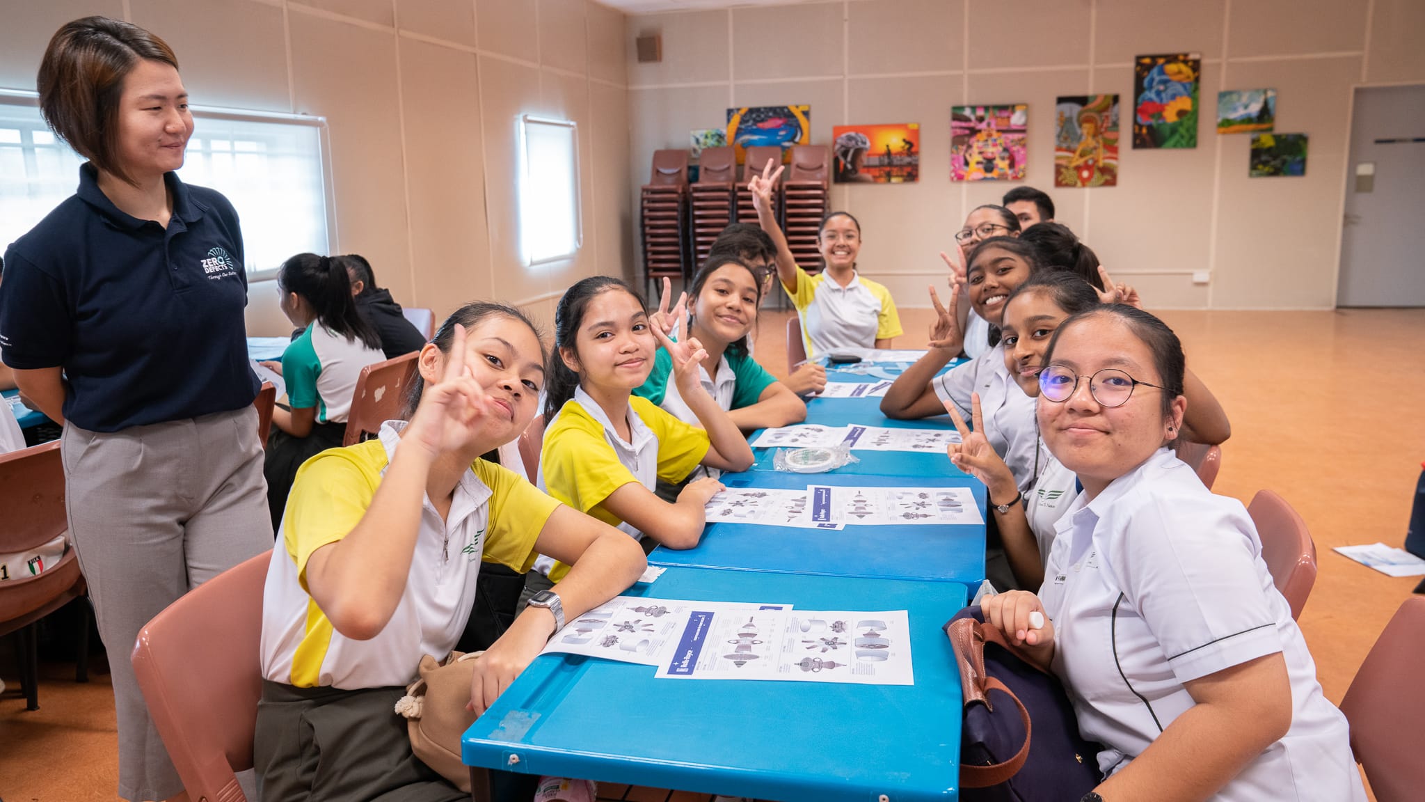 Students in school uniforms sit at a blue table making peace signs, with a teacher standing nearby in a classroom.